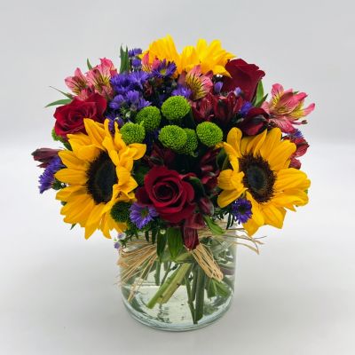 Reminiscent of strolling through a field of flowers, this colorful arrangement captures features magnificent sunflowers, alstroemeria and poms set in a trendy mason jar with a natural raffia bow.